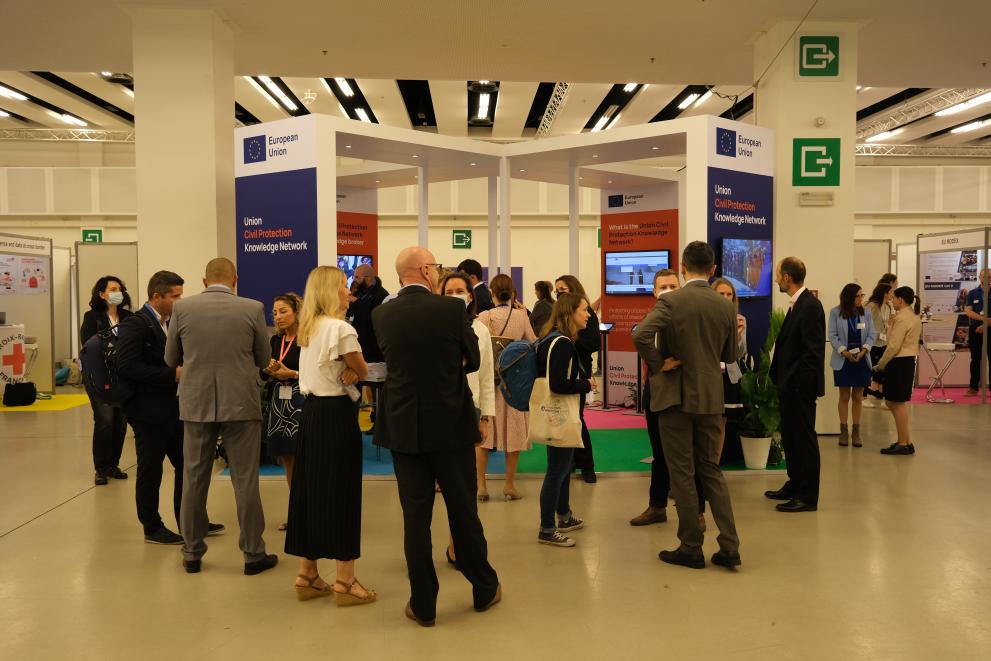  A group of around 20 people visit an exhibition stand at a conference. The exhibition stand is in the shape of a parge cross and is open inside. It has the European Union emblem and the panels are titled Knowledge Network. 