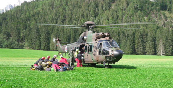 A cave search and rescue team are beside a helicopter in an open field with forest behind. They have disembarked from the helicopter and are preparing their cave rescue equipment.