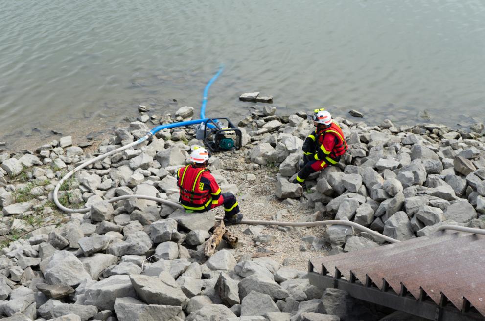 Two emergency services personnel sit on rocks beside a river. They are operating pumps to reduce water levels.