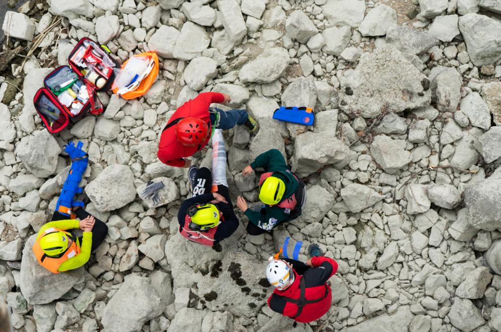 A group of emergency rescuers sit on rocks treating a patient with an injured leg. Their medical kit is beside them.