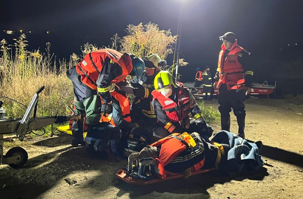A search and rescue team work at night, grouped around a flood victim who is strapped into an evacuation harness ready to be moved to receive additional medical care.jpg