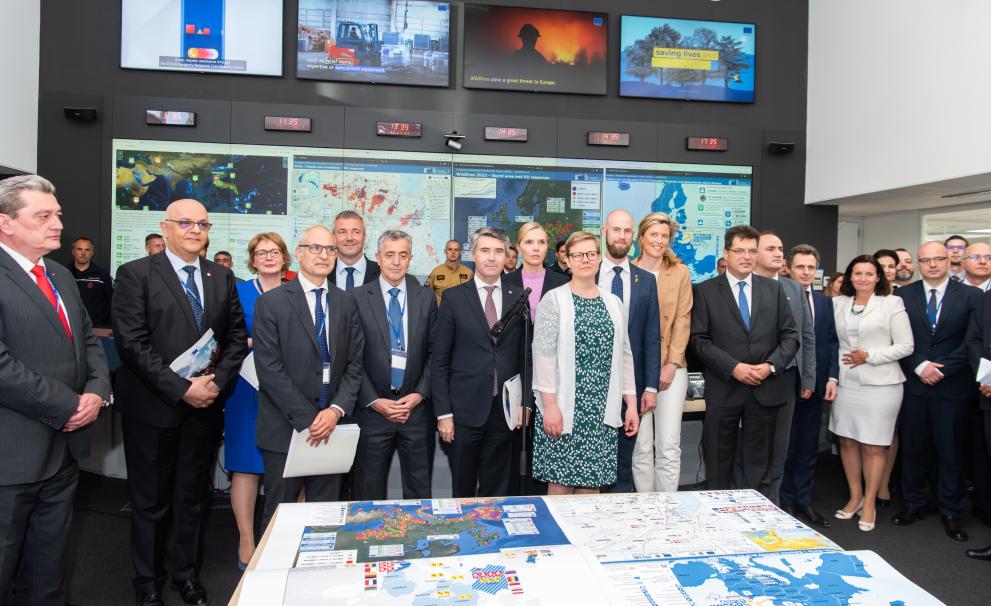 A group of people stand behind a table displaying maps. Behind them are screens showing weather systems and news channels.