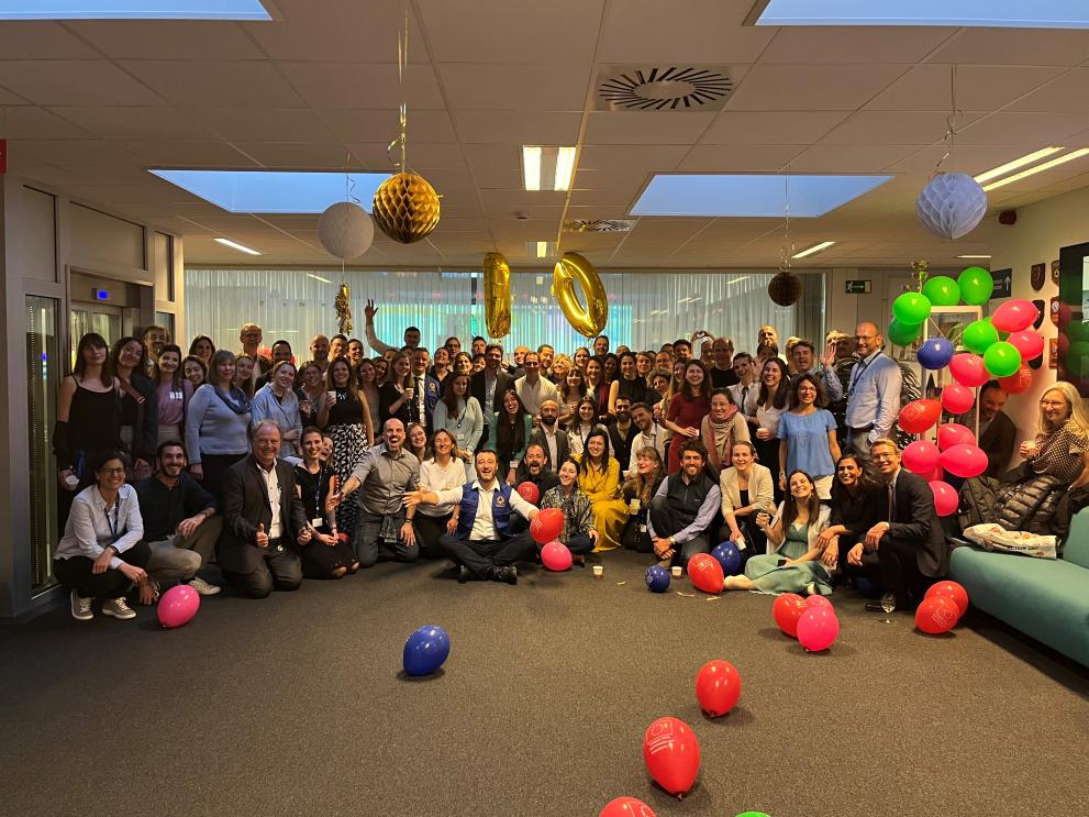 A group of people sit and stand in a large operations room. There are red balloons in front of them.