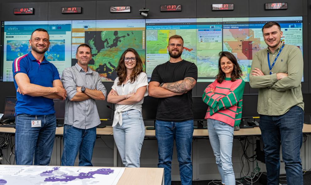 A group of people stand smiling with their arms folder. Behind them are screens with maps