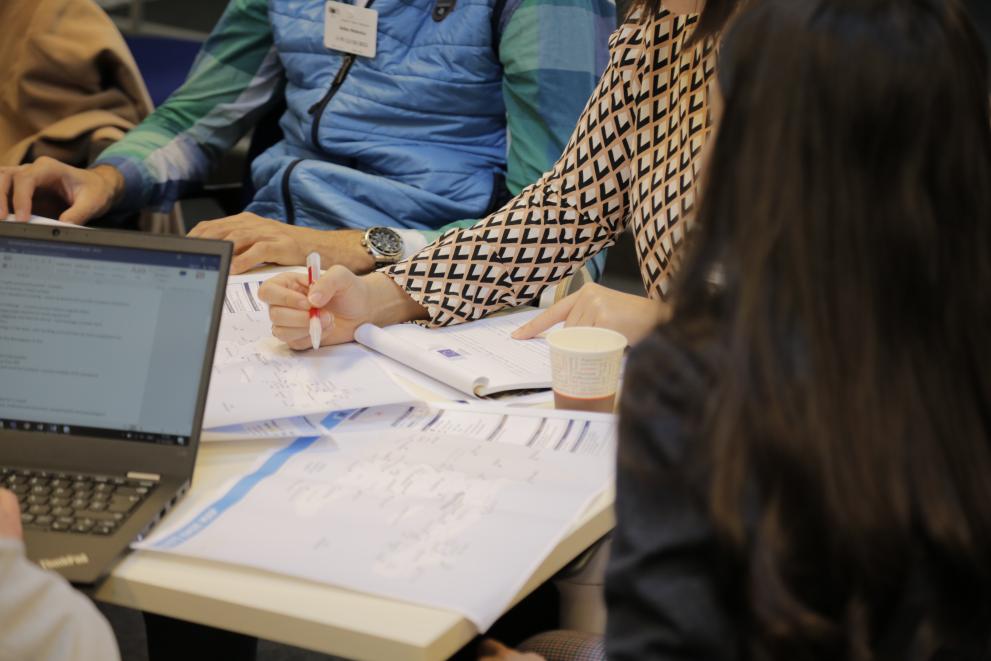 Close-up of persons around a table, one writing on a document, another at a laptop.