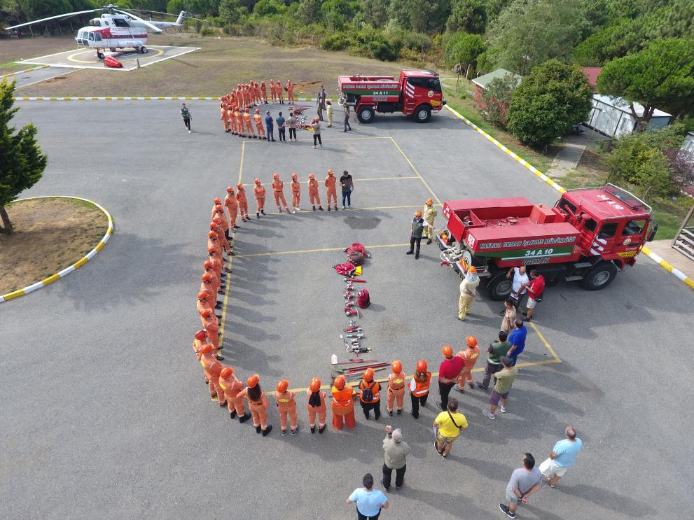 A parking area with a white and red helicopter at the far end and, closer to the camera, two firetrucks. In front of each firetruck there is a group of people (most dressed in orange overalls and hard hats) in a half circle listening to explanations given by a person in front of them. There is also firefighting equipment spread out for display on the ground in the middle of each half circle.