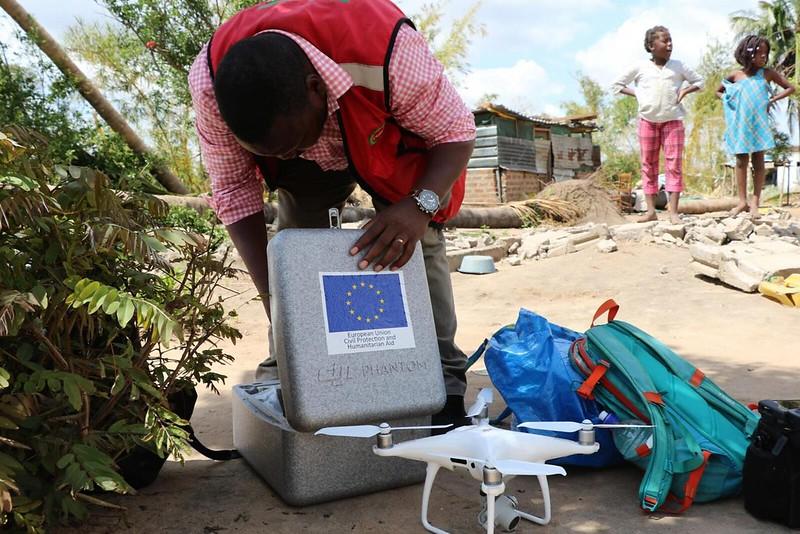 A person opening a box with the EU logo for Civil Protection and Humanitarian Aid in a sandy area. In front of the box, there is a drone waiting on standby.