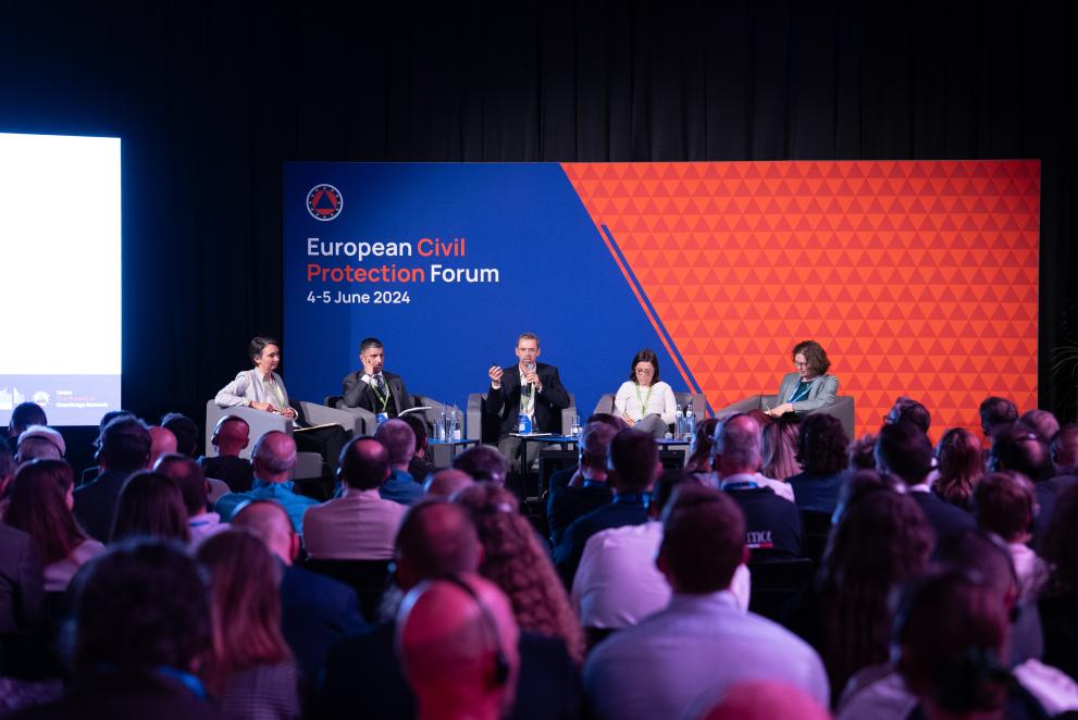 panel during the civil protection forum 2024, four panelist are on stage, orange and blue backdrop, man in the middle is speaking.