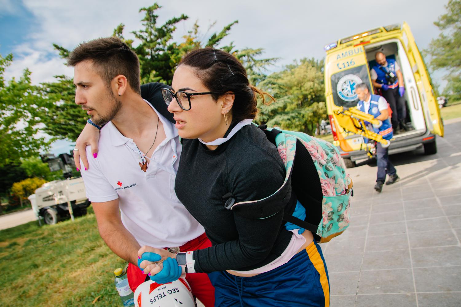A male medical staff member supports a woman. An ambulance is behind them.