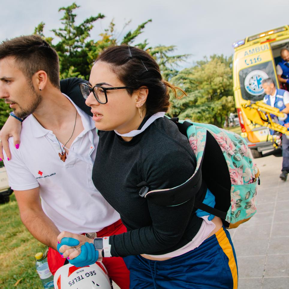 A male medical staff member supports a woman. An ambulance is behind them. 