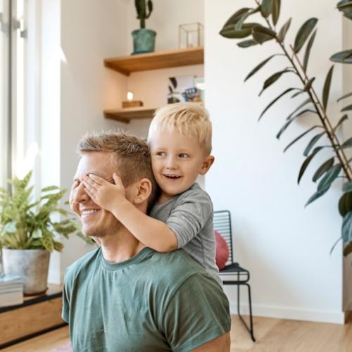 A young boy with blond hair covers his father's eyes. Both are laughing.