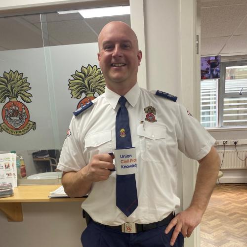 A man in firefighter uniform stands in an office. He is holding a mug and smiling at the camera.