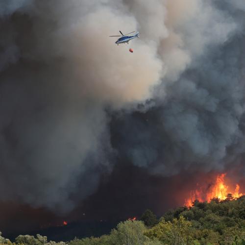 A helicopter with water bombing equipment flies above a heavily forested hillside that is on fire.