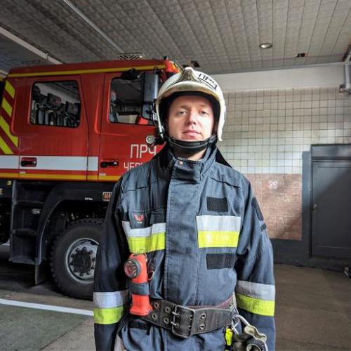 Ukrainian firefighter Andriy stands in front of a fire engine. He is dressed in firefighting protective clothing.