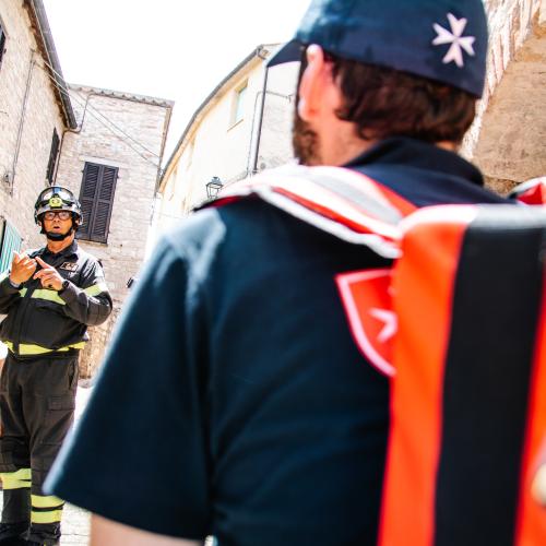 A member of the emergency medical team wearing a medical kit bag is speaking with a fire service personnel member.