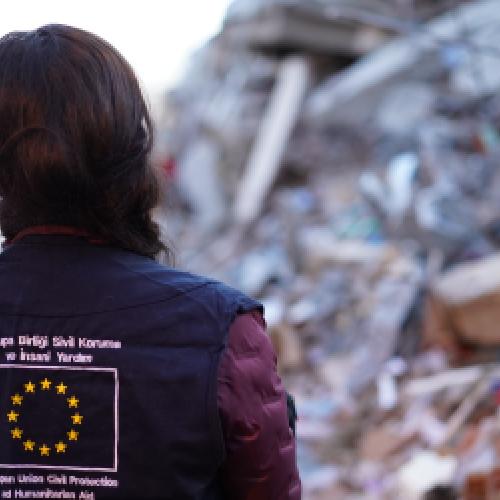 A person wearing an EU civil protection vest stands in front of a destroyed building