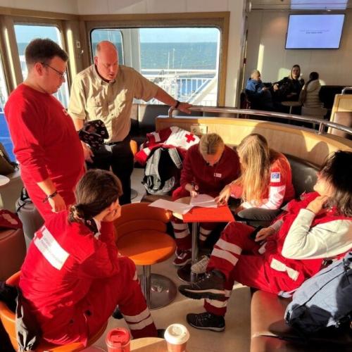 Persons wearing red shirts or vests sitting and standing around a small coffee table inside what appears to be a boat. 