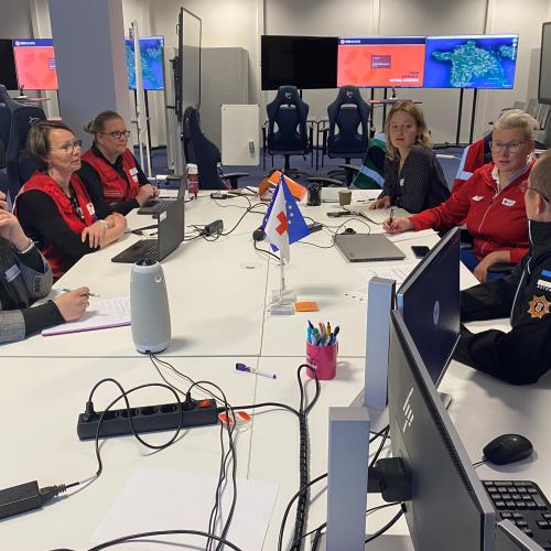 Women sitting at a table discussing, with large computer screens in the background.