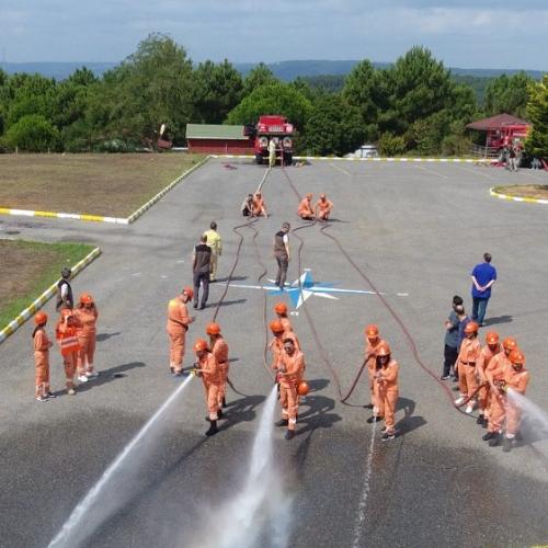 In a large cemented area, persons dressed in orange suits and hard hats spraying water from four fire extinguishing hoses, with other persons observing or helping.