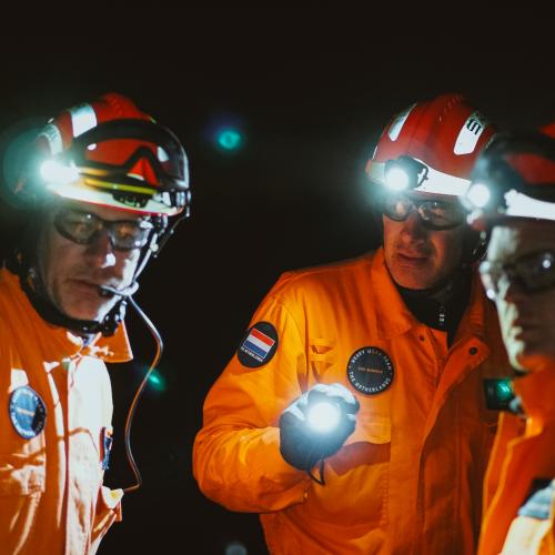Three rescuers stand together. They carry torches and are wearing red hard hats and orange safety suits..