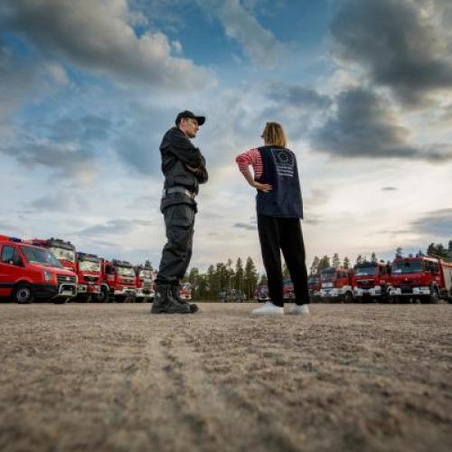 Two persons in uniform standing in an open outdoor area with several firetrucks in the distance.
