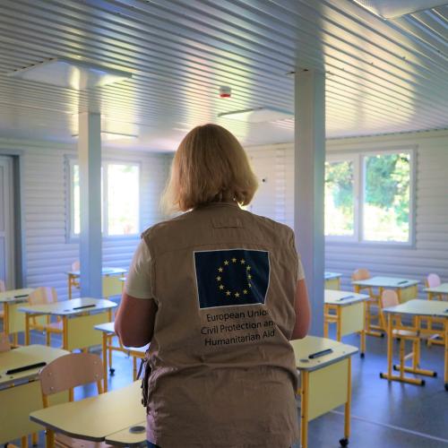 Woman wearing a EU vest stands in front of an empty classroom.