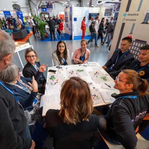 A group of participants during an interactive session at the 8th Civil Protection Forum. Most participants are seated. A woman is explaining something to the group.