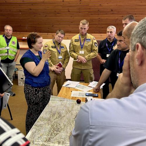 Participants of the ModTTX5 attend a meeting, standing infront of a map of the affected area.