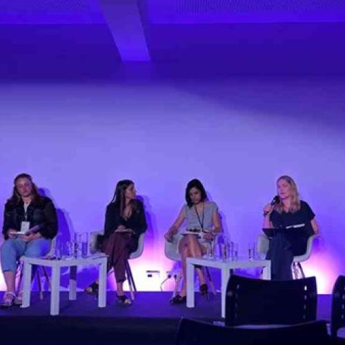 Photograph of a panel discussion with six people seated on stage under purple lighting, each with microphones and water glasses on small tables in front of them. The setting suggests a formal event or conference focused on dialogue or presentations, with audience chairs visible in the foreground.