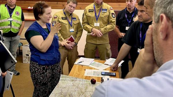 Participants of the ModTTX5 attend a meeting, standing infront of a map of the affected area.