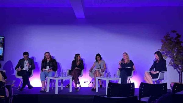 Photograph of a panel discussion with six people seated on stage under purple lighting, each with microphones and water glasses on small tables in front of them. The setting suggests a formal event or conference focused on dialogue or presentations, with audience chairs visible in the foreground.